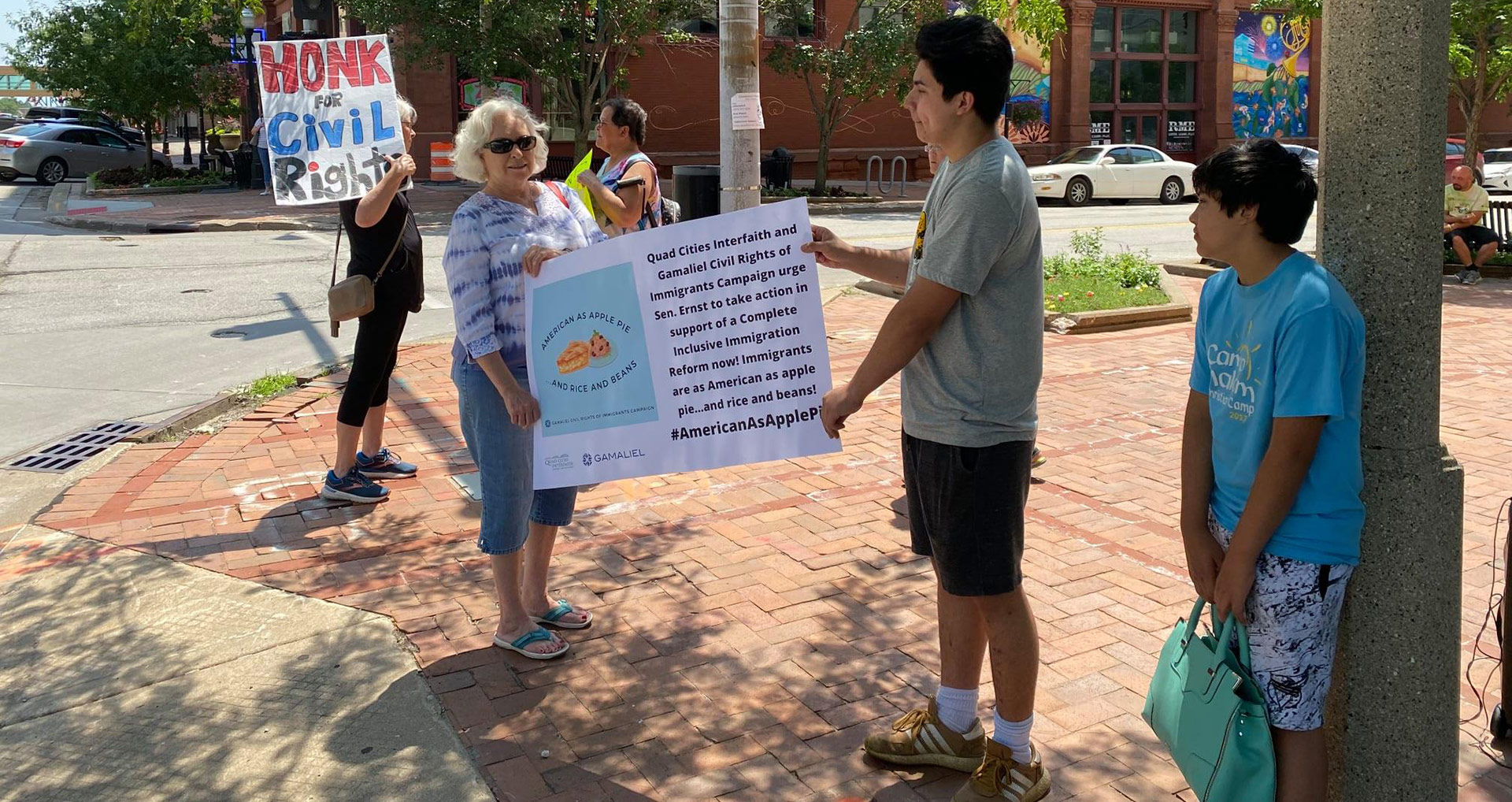 Several people stand outside under trees next to an intersection holding signs supporting civil rights