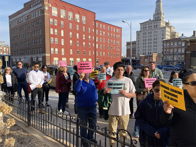 A gathering of protestors holding signs in English and Spanish expressing support for excluded and essential workers